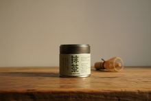 Load image into Gallery viewer, A tin of Uji Matcha tea sits on a rustic wooden cutting board against a textured gray wall. The cylindrical tin has a dark gray lid and a light gray-green label with black Japanese characters and English text reading 'UJI MATCHA'.