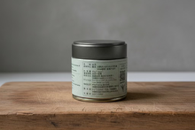 Load image into Gallery viewer, A small round jar of matcha green tea from Doti Farm, featuring a dark gray lid and a beige label with black text, placed on a wooden surface against a textured gray wall.