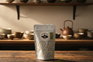 A stand-up pouch of Chagama tea on a wooden surface against a light gray wall. The pouch is white with a circular black and white Chagama logo and a green vertical label with Japanese characters.